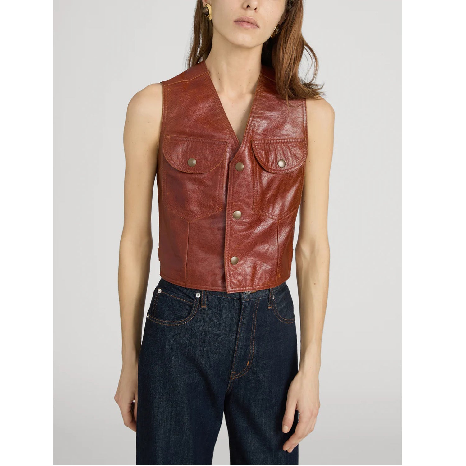 A model wearing a cropped brown leather vest with front button closures and flap pockets, paired with dark blue jeans, against a light gray studio background.