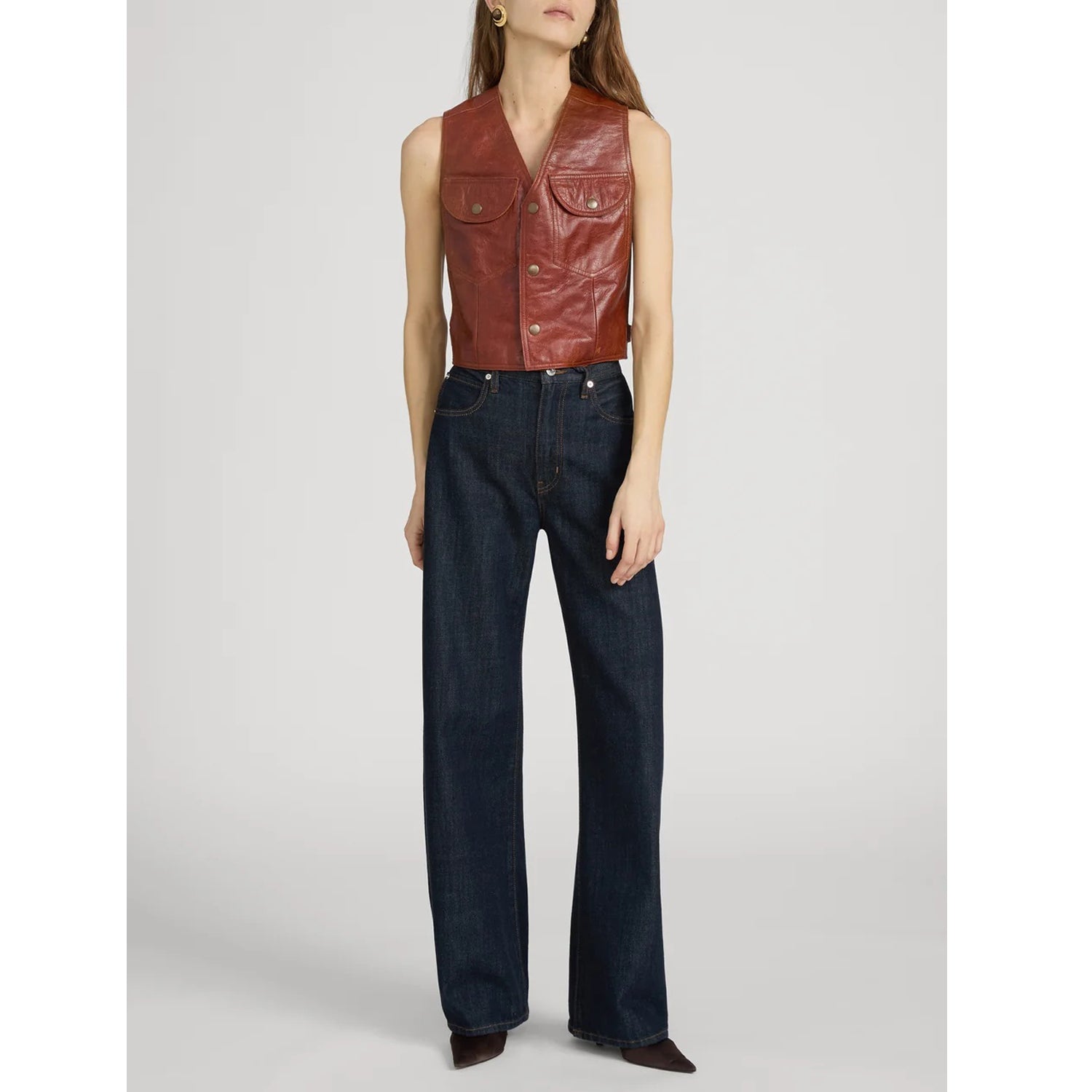 A model wearing a rust-brown leather sleeveless vest with button details and dark blue wide-leg jeans, standing against a light gray studio background.