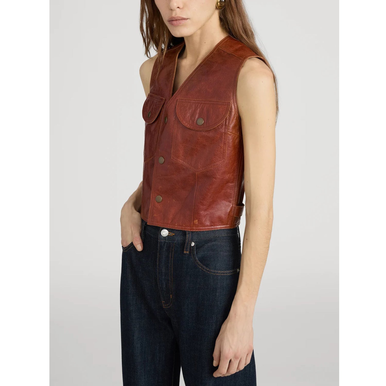 A model wearing a brown leather sleeveless vest with front flap pockets and dark blue jeans, photographed against a light gray studio background.